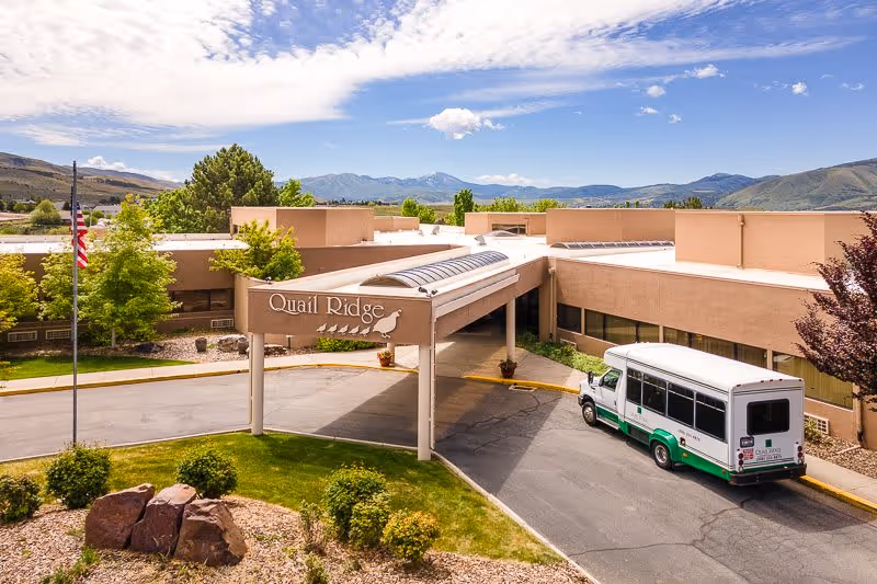 Exterior view of Quail Ridge senior living facility with a covered entrance, a shuttle bus parked nearby, landscaped greenery, and mountains in the background under a partly cloudy sky.
