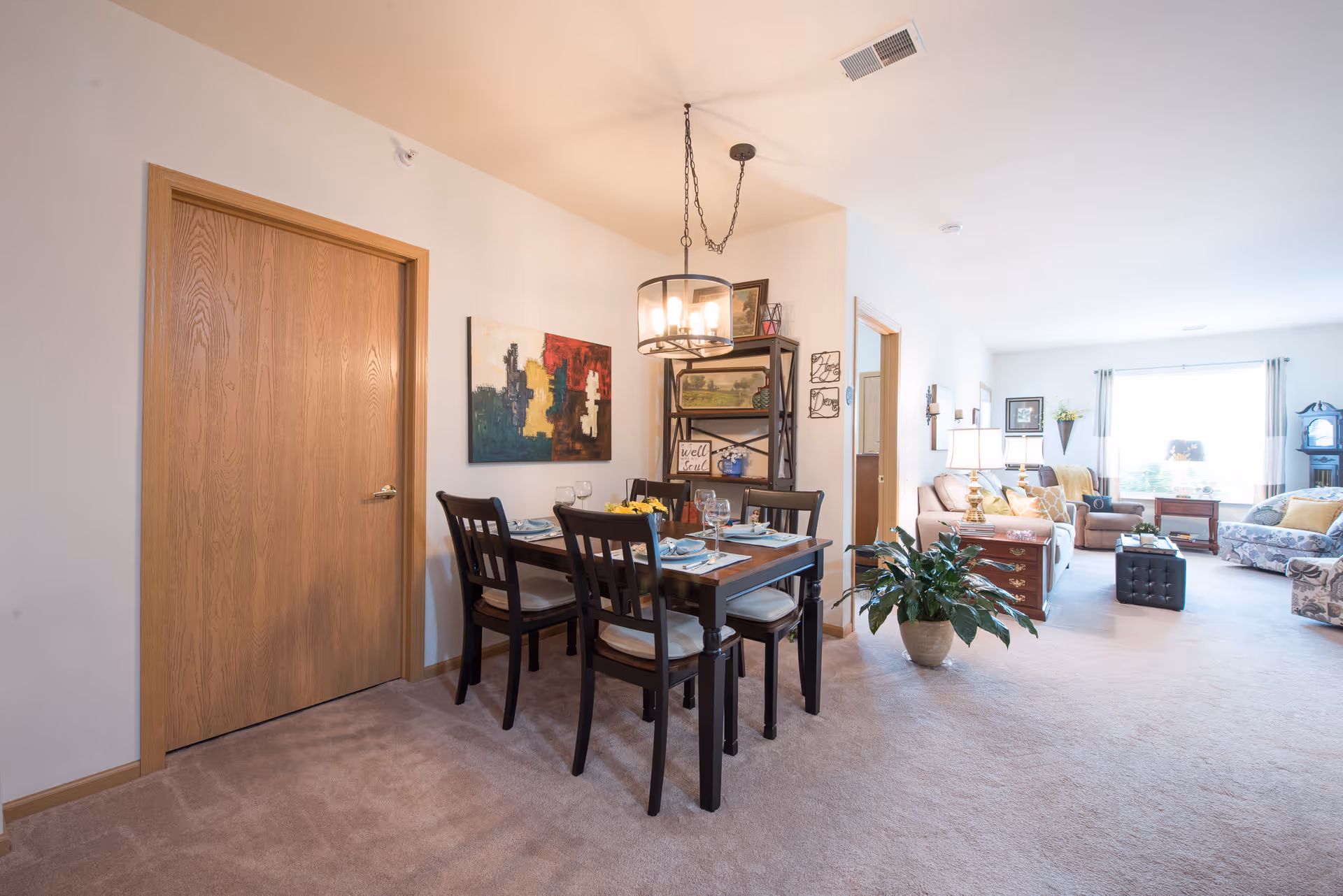 A bright and spacious interior featuring a dining area with a wooden table set for four, a modern chandelier above, and a colorful abstract painting on the wall. Adjacent to the dining area is a cozy living room with multiple sofas, a large window with curtains, a side table with lamps, and a potted plant on the carpeted floor.