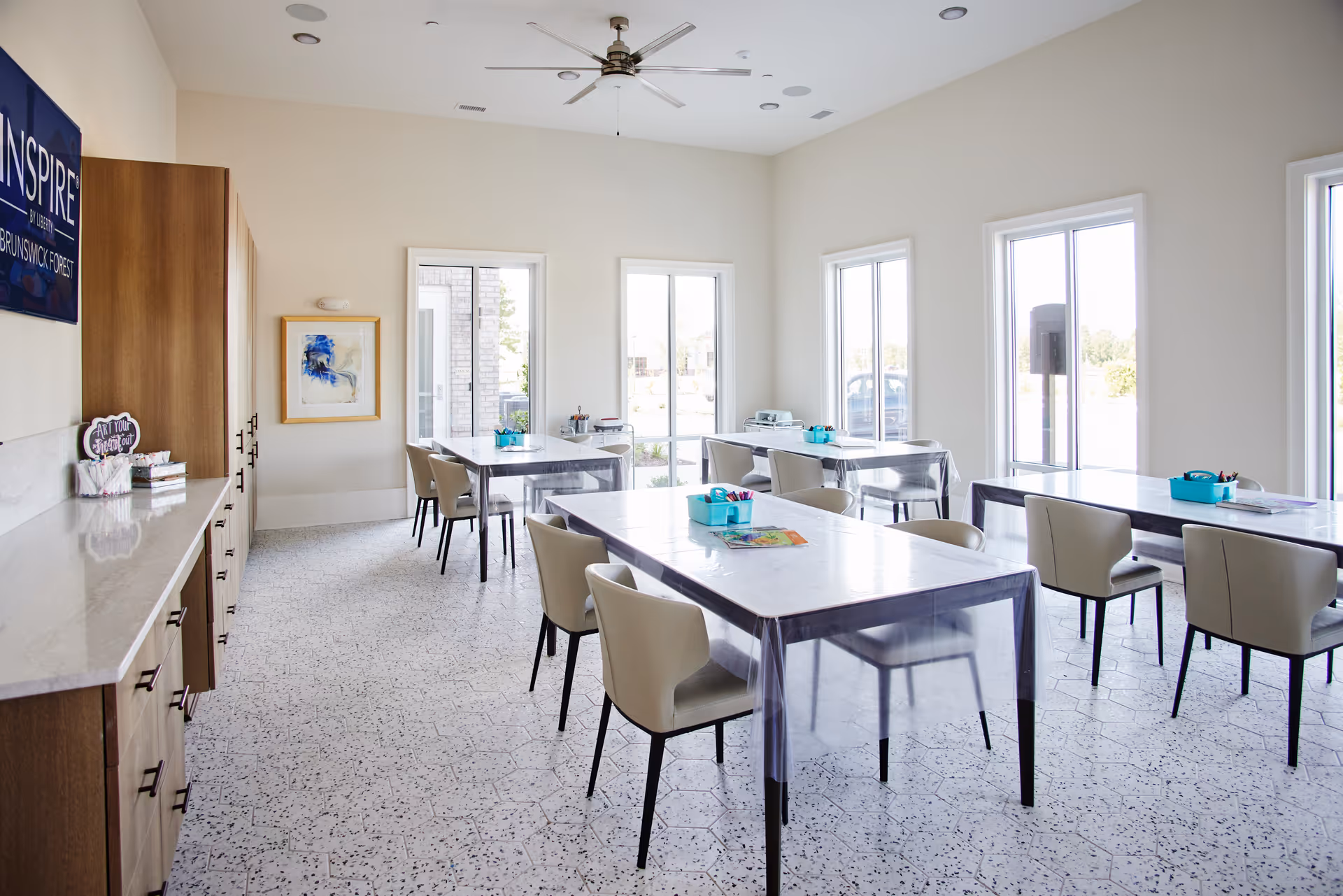 Bright room with several tables covered in clear plastic, each surrounded by beige chairs. The room has large windows letting in natural light, a ceiling fan, and a counter with cabinets along one wall. Art supplies and a sign that says 'Art Your Heart Out' are visible on the counter.