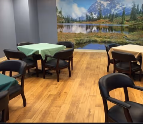 Small dining area with round tables covered in pastel tablecloths, dark chairs on a wood floor and a large mountain-lake mural on the wall.