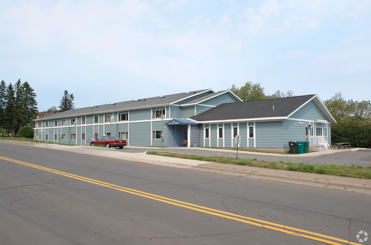 Exterior view of a two-story blue building with white trim, identified as Lakeland Shores Apartments. The building has multiple windows, a small entrance canopy, a parked red car, and is situated next to a paved road with yellow dividing lines. There are trees and greenery in the background under a partly cloudy sky.