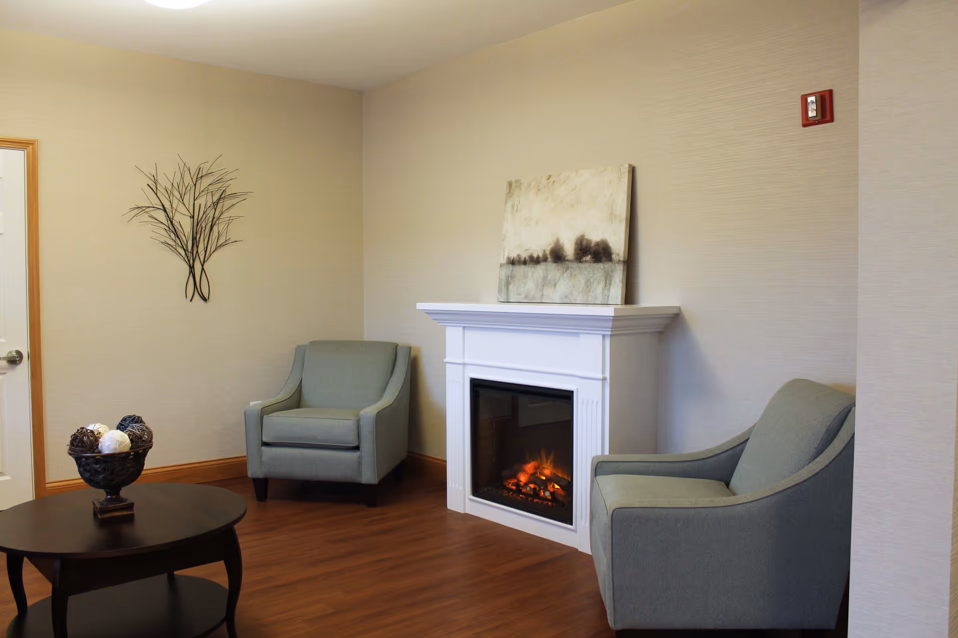 A cozy sitting area with two gray armchairs positioned near a white electric fireplace. Above the fireplace is a landscape painting. A round dark wooden coffee table with decorative balls in a bowl is in the foreground. The walls are beige with a metal wall art piece shaped like branches on one side.