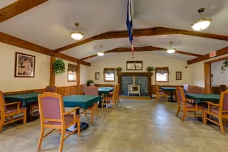 Community dining room with multiple tables and chairs, exposed wooden ceiling beams, a flag hanging from the ceiling, and a fireplace on the far wall.