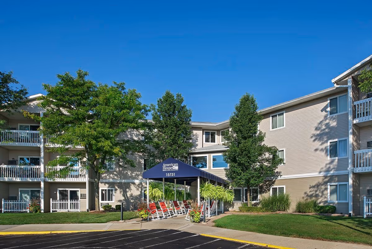 Exterior view of a three-story senior living facility building with beige siding and white balconies. There is a covered entrance with a blue canopy labeled 'American House 15731' and several red rocking chairs underneath. The area is landscaped with green trees, bushes, and a well-maintained lawn under a clear blue sky.