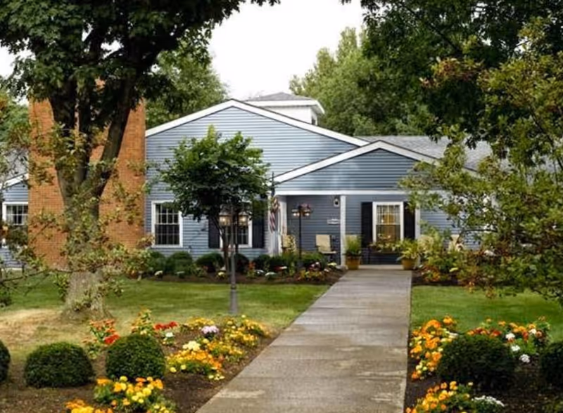 Front exterior view of a single-story blue building with white trim, surrounded by green trees, bushes, and colorful flower beds. A concrete pathway leads to the entrance, which has two chairs and potted plants on the porch.