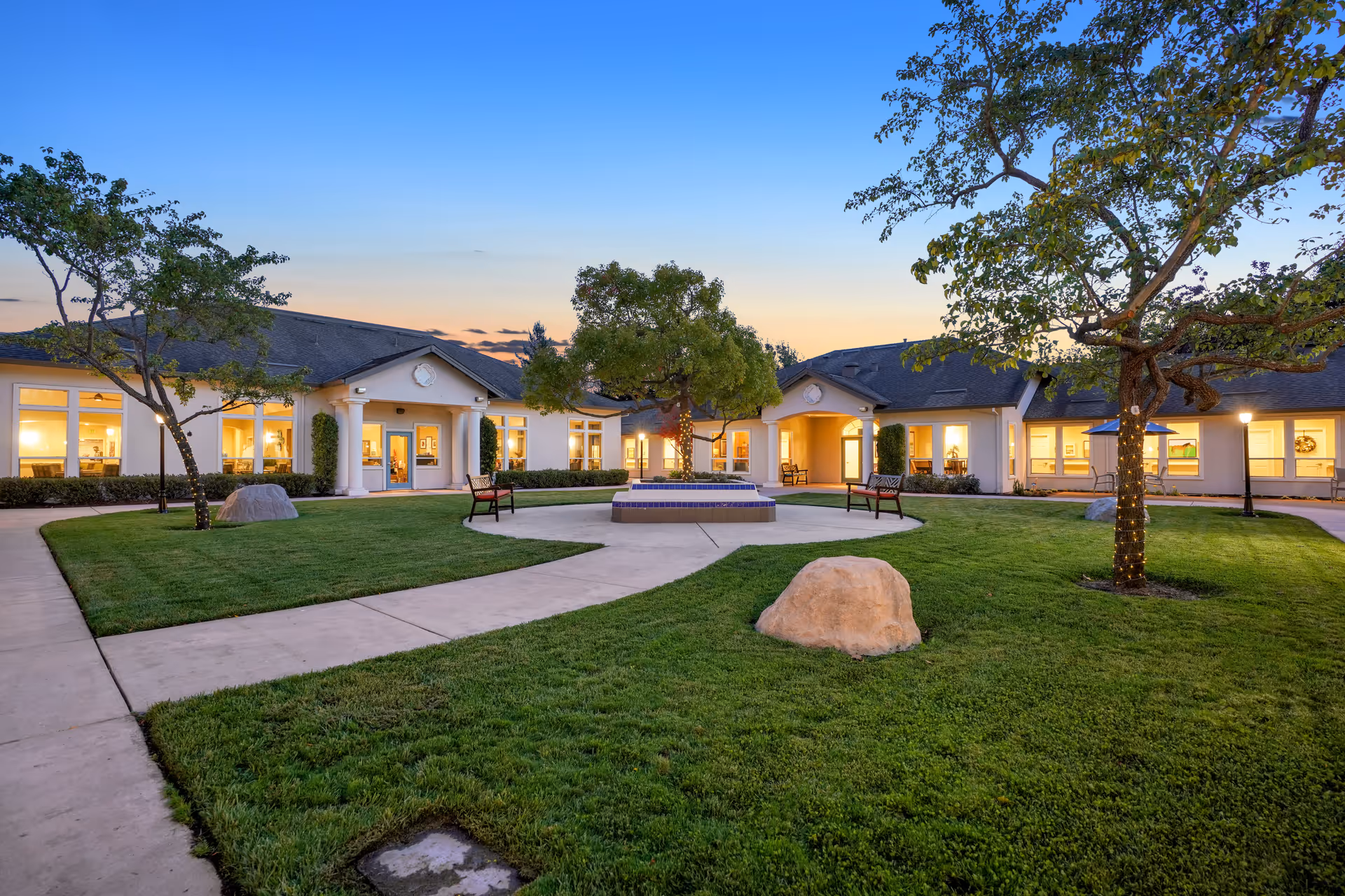 Outdoor courtyard area of a senior living facility at dusk with well-maintained green grass, trees wrapped with string lights, benches, a central fountain, and a single-story building with large windows and warm interior lighting.