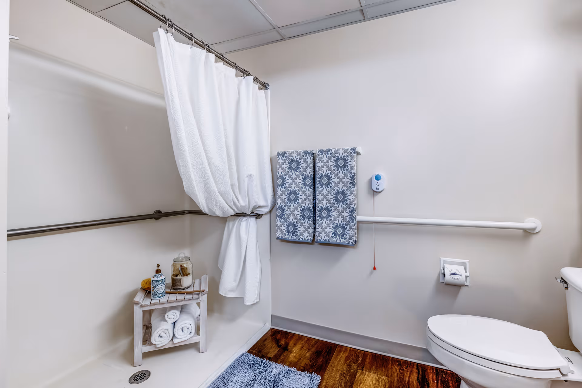 A bathroom with a white shower curtain partially drawn to the side, a small wooden shelf holding rolled white towels and bath items, two patterned blue and white towels hanging on a wall-mounted bar, a white toilet, and a wooden floor with a blue bath mat. The walls are light-colored and there are grab bars installed for accessibility.