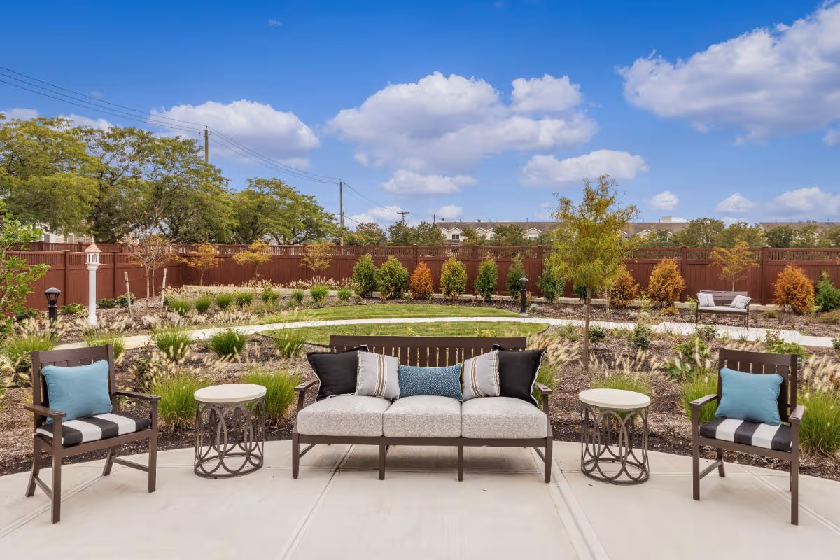 Outdoor patio area with a cushioned sofa and two armchairs, each with decorative pillows, arranged around two small round tables. The patio overlooks a landscaped garden with various plants, shrubs, and a wooden fence under a blue sky with scattered clouds.