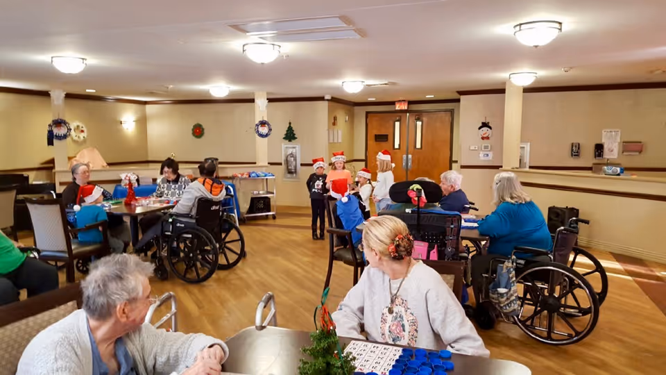 A group of elderly people, some in wheelchairs, sitting around tables in a large room decorated with holiday wreaths. Several children wearing Santa hats are standing and interacting with the seniors. The room has wooden flooring and beige walls with ceiling lights.