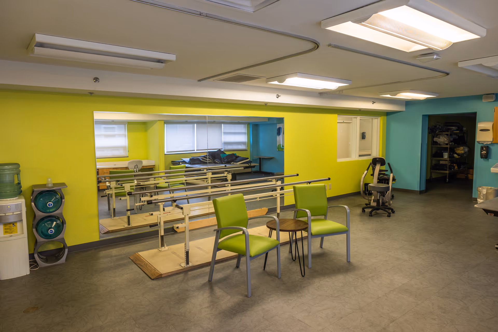 Rehabilitation room with parallel walking bars, green chairs, a water cooler, and a wall-length mirror.