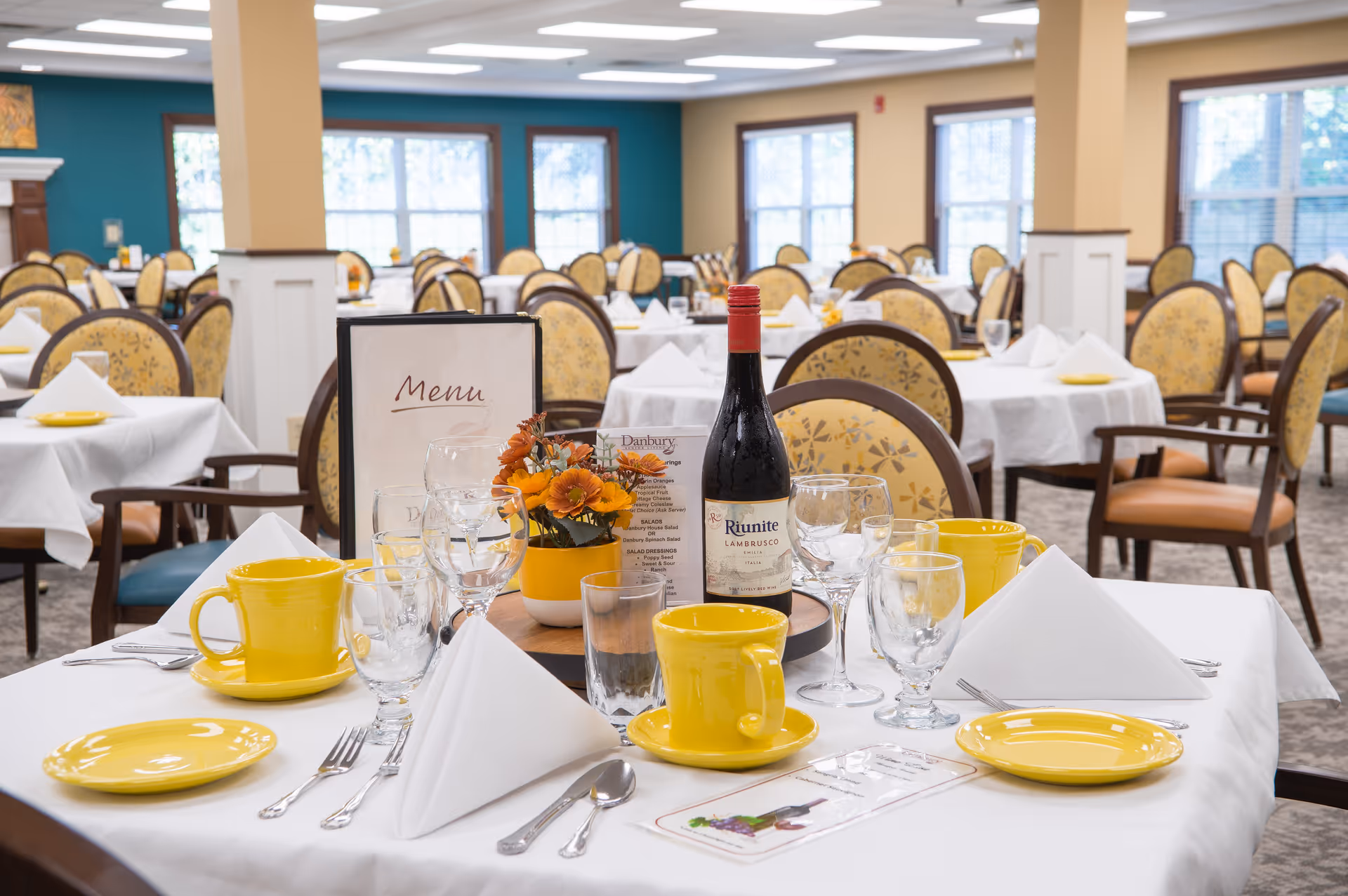 A dining room in a senior living facility with tables covered in white tablecloths, set with yellow plates, yellow mugs, glassware, silverware, and white folded napkins. A bottle of red wine and a small floral arrangement are placed on the table in the foreground. The room has multiple chairs with floral upholstery and large windows letting in natural light.