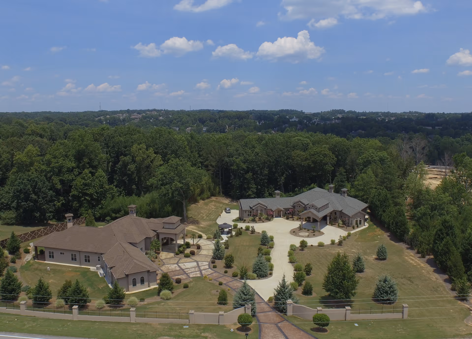Aerial view of a senior living facility named Angels Garden Senior Community PCH Assisted Living & Memory Care, showing two large buildings surrounded by landscaped grounds with trees, shrubs, and a driveway. The facility is set in a green, wooded area under a partly cloudy blue sky.