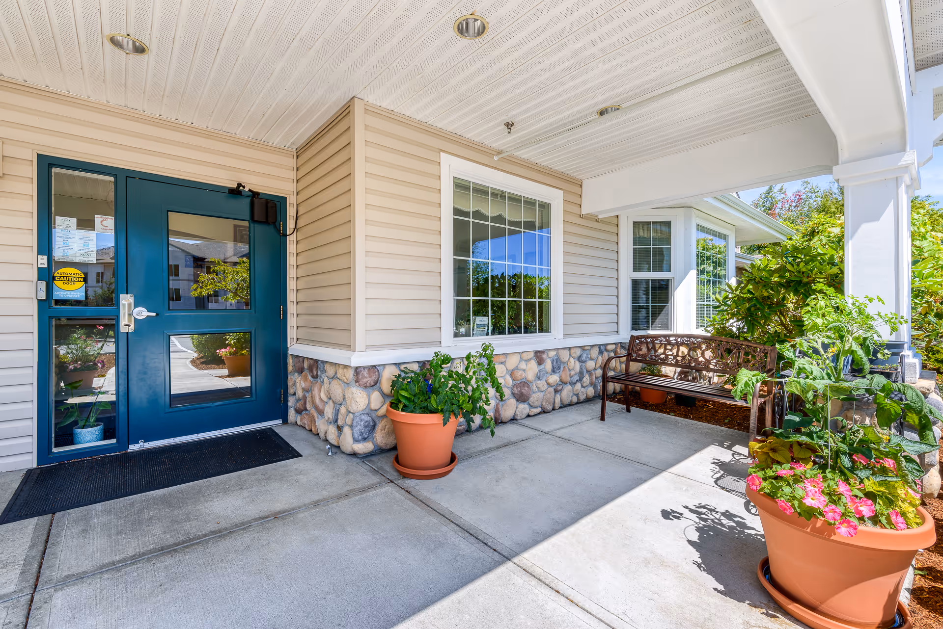 Covered building entrance with a blue door, stone-accented siding, potted plants and a bench on the porch.