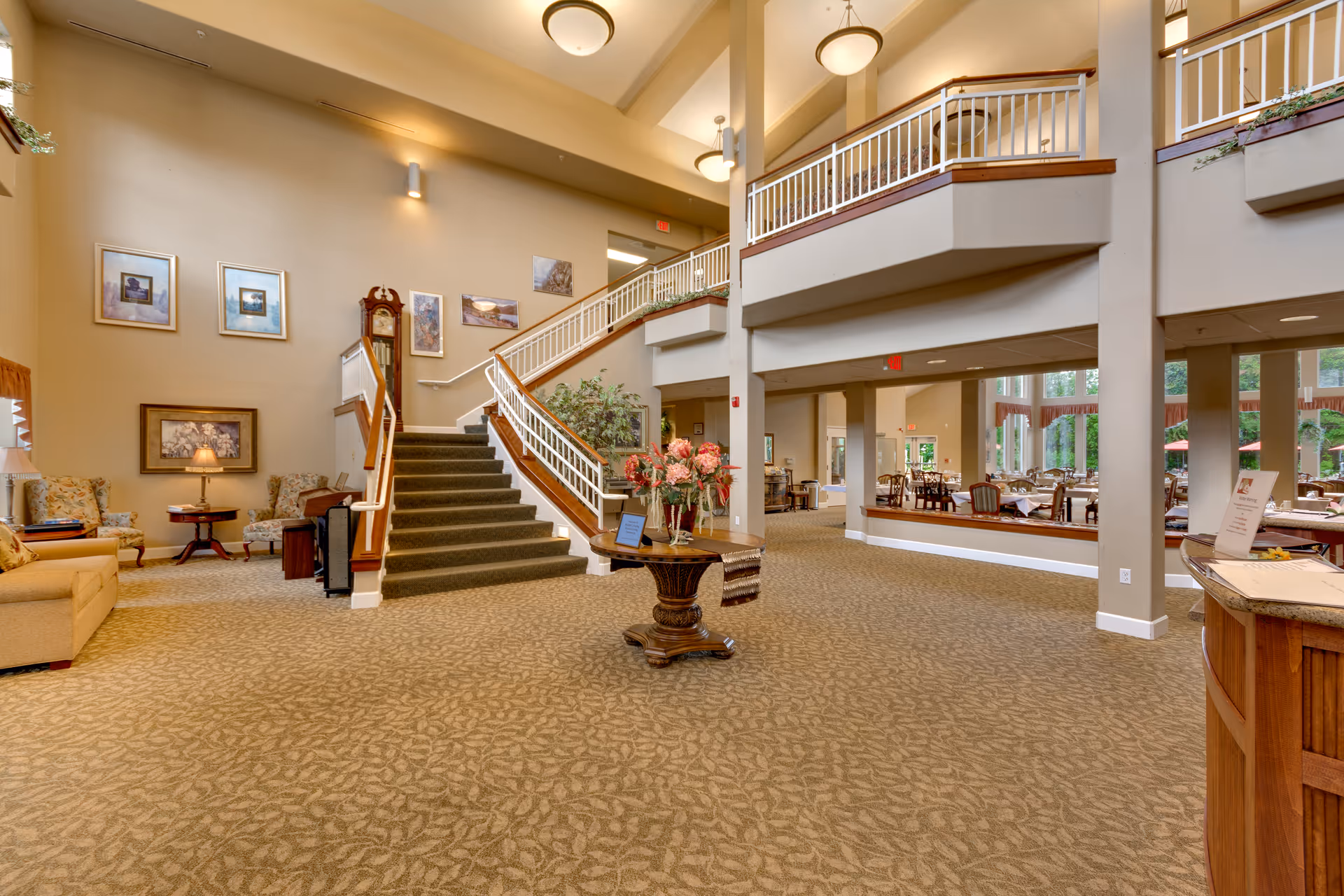 Spacious interior of a senior living facility with a carpeted floor, a central wooden table with a floral arrangement, a staircase with white railings leading to an upper level, and a seating area with armchairs and a lamp. The dining area with tables and chairs is visible in the background through large windows.