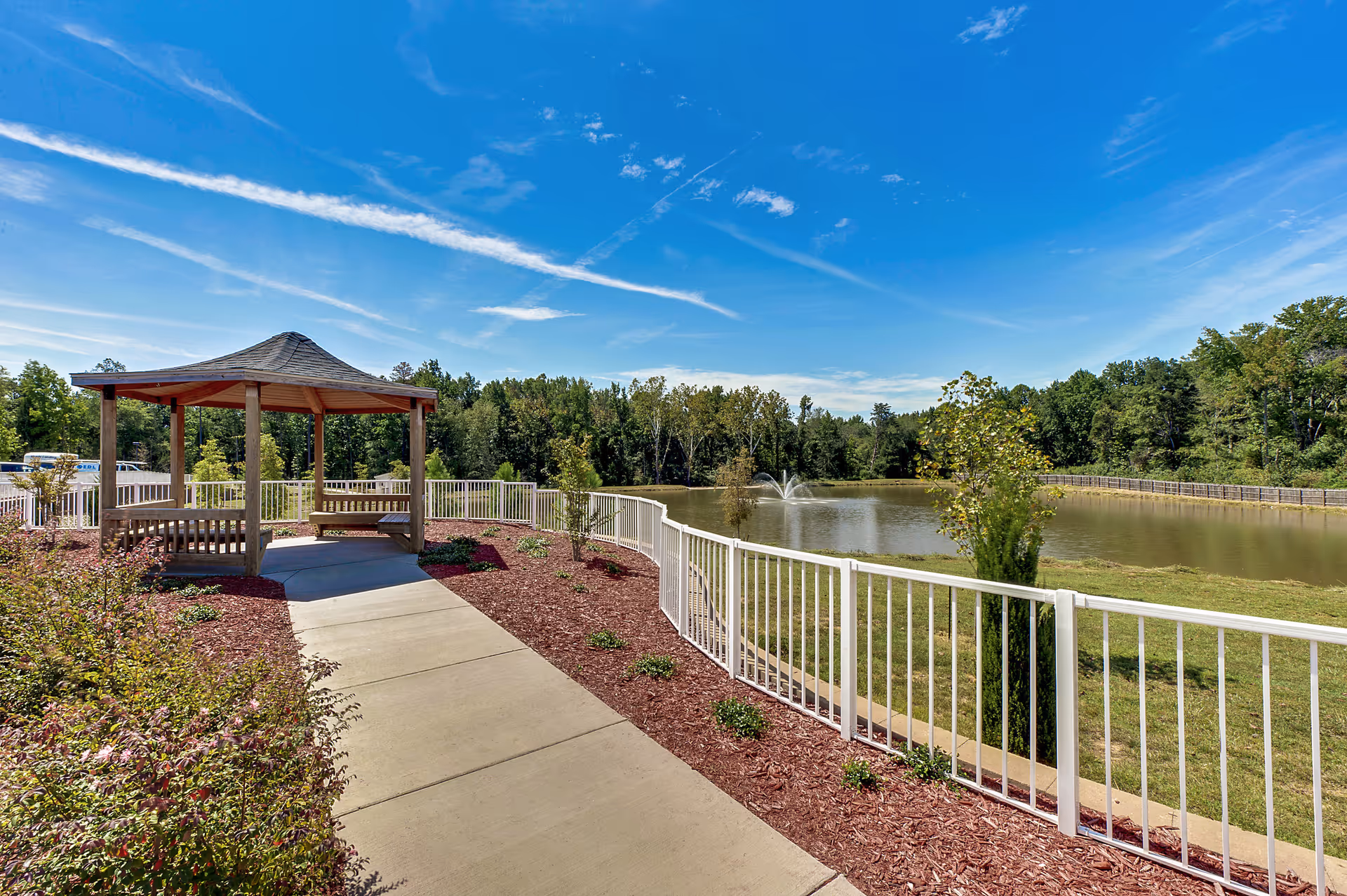 Gazebo-covered seating area beside a paved walkway overlooking a fenced pond with a fountain and trees under a blue sky.