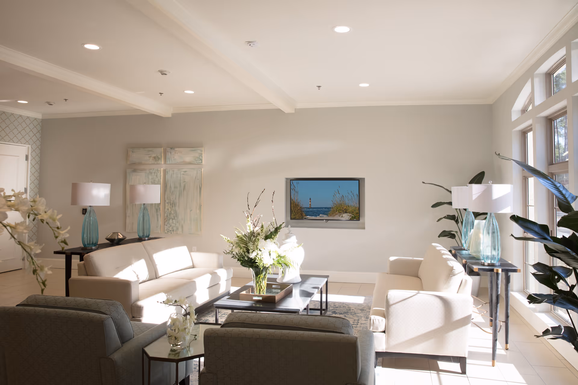 A bright and airy living room with two beige sofas, two armchairs, a coffee table with a floral arrangement, side tables with blue glass lamps, large windows letting in natural light, and a wall-mounted TV displaying a lighthouse scene.