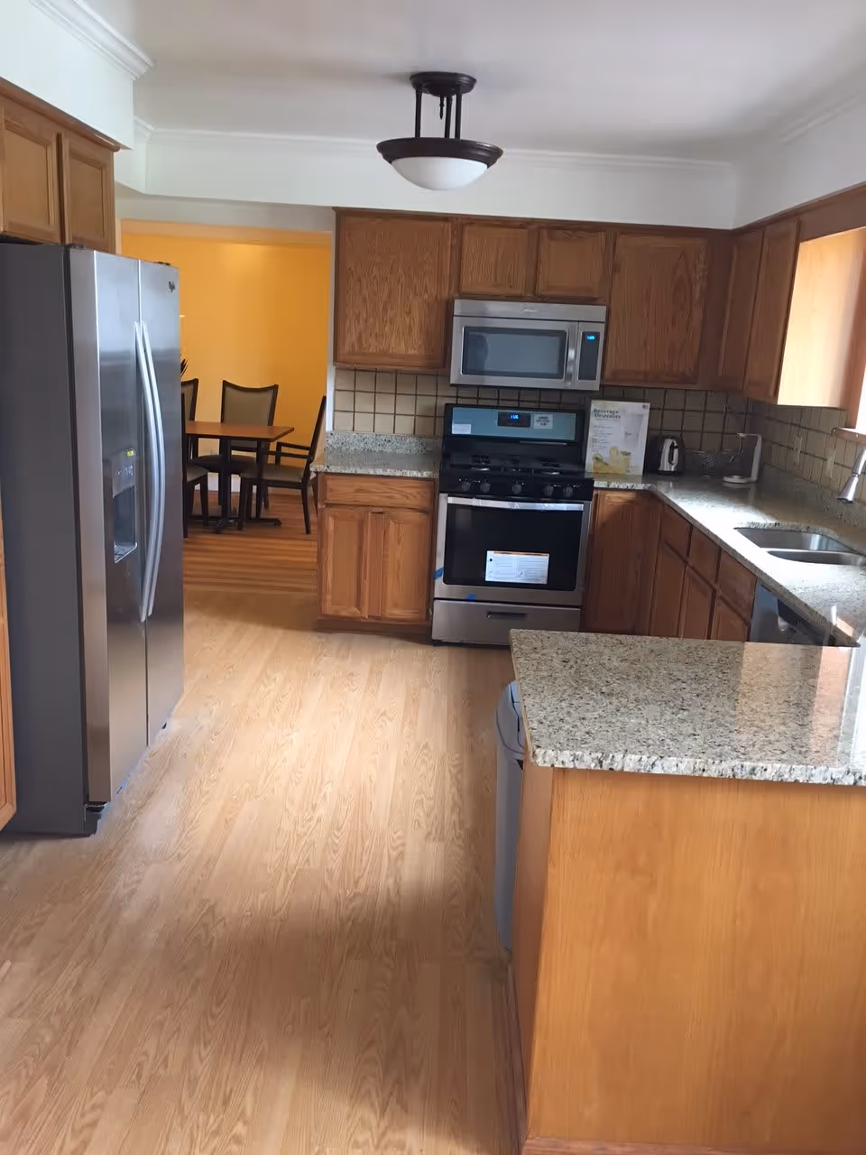 Bright kitchen with wooden cabinets, granite countertops, a stainless steel refrigerator, stove and microwave, and a dining area visible through a doorway.