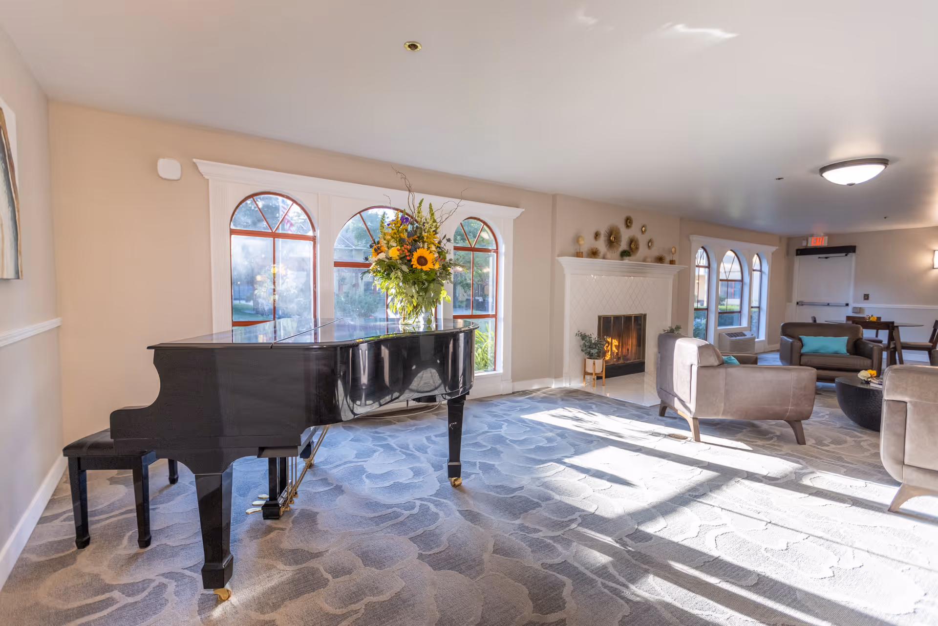 A bright and spacious living room area with a black grand piano adorned with a large bouquet of flowers on top. The room features large arched windows letting in natural light, a lit fireplace with decorative wall art above it, and several comfortable armchairs arranged around a coffee table. The carpet has a subtle floral pattern, and the walls are painted a light beige color.