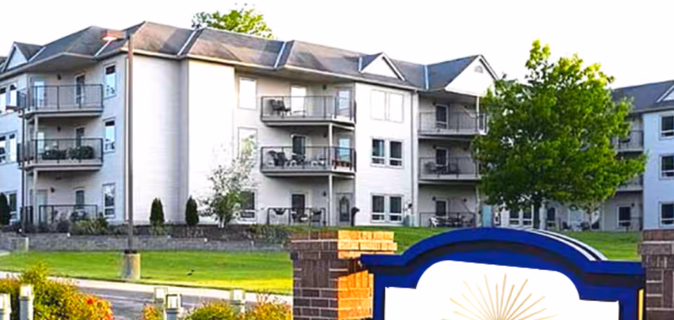 Exterior view of a multi-story senior living building with balconies, lawn, and a partial entrance sign in front.