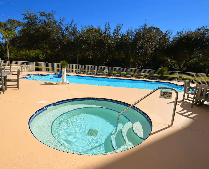 Outdoor pool area at the Landing Of St Andrew featuring a circular hot tub with steps and a metal handrail in the foreground, a larger rectangular swimming pool with lounge chairs along the fence in the background, and trees beyond the fence under a clear blue sky.