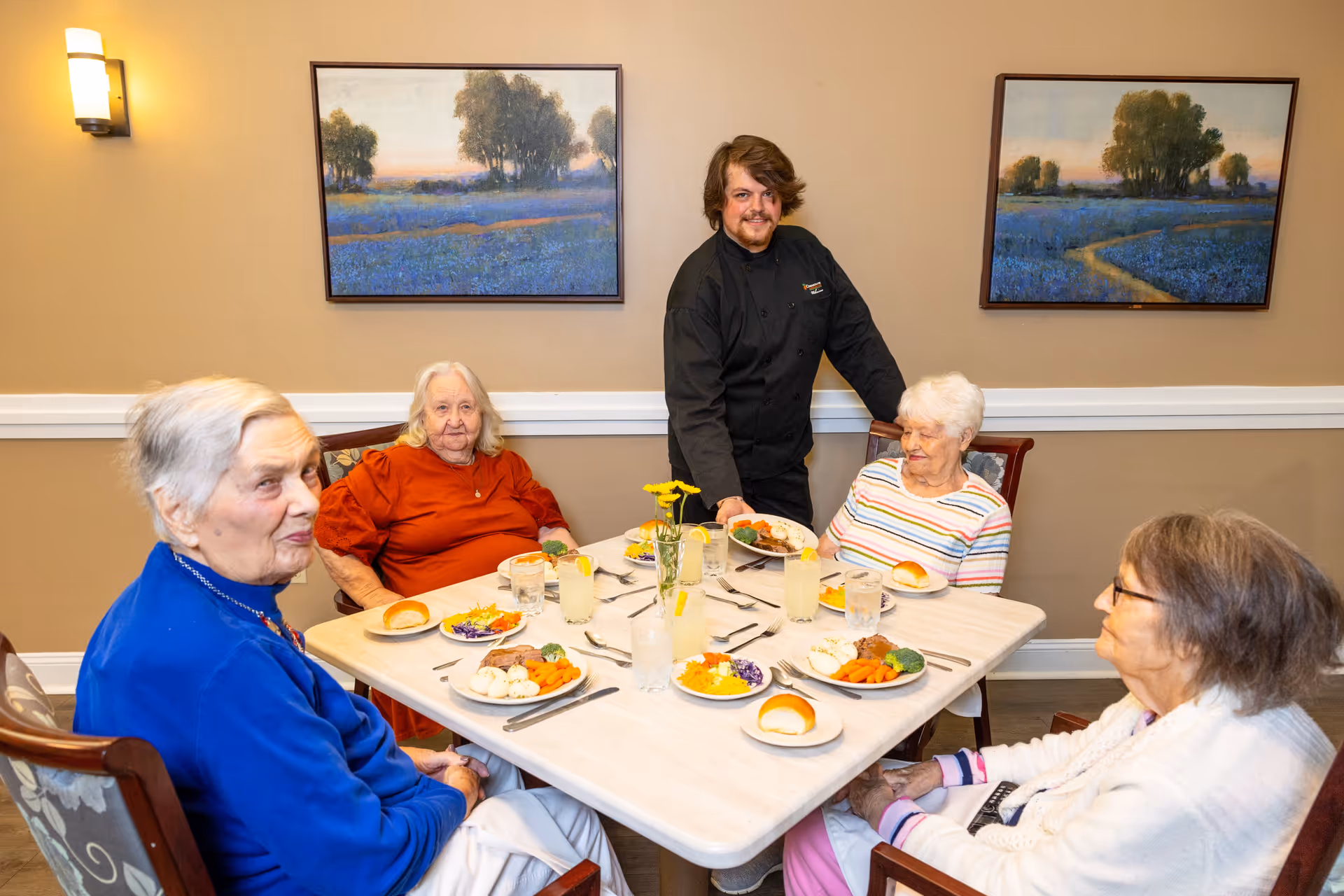 A group of four elderly women seated around a dining table with plates of food and glasses of lemonade. A man in a black chef's uniform is standing and serving food to one of the women. The room has beige walls with two landscape paintings and a wall sconce light.