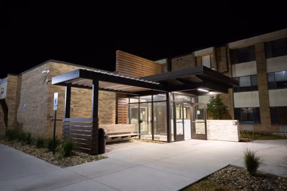 Night view of the entrance to Keystone Ridge Post Acute Nursing and Rehabilitation facility, featuring a modern covered walkway with wooden slats, glass doors, and exterior lighting illuminating the pathway and building facade.