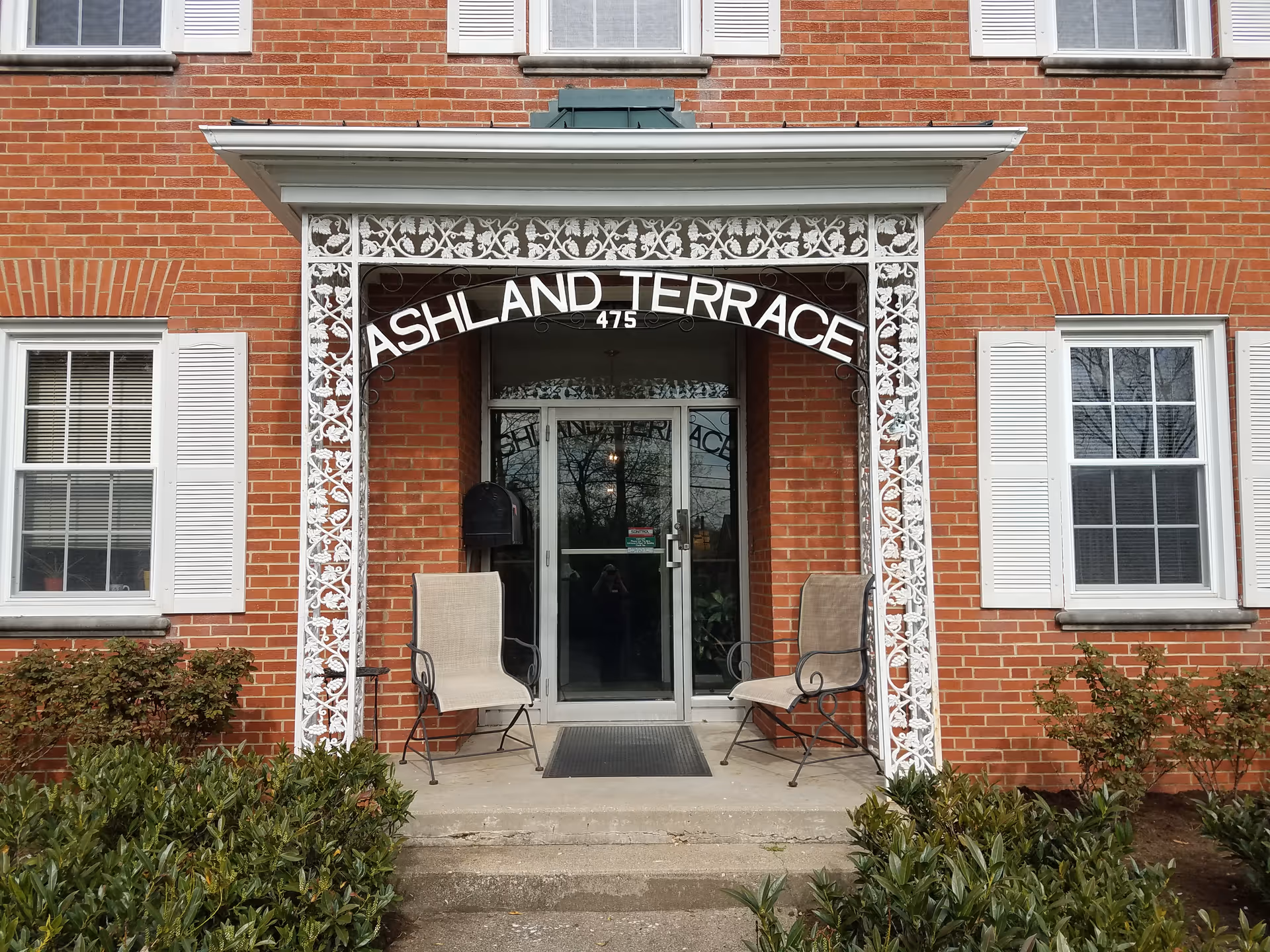 Brick building entrance with a white decorative awning reading ASHLAND TERRACE and two chairs flanking the doorway.