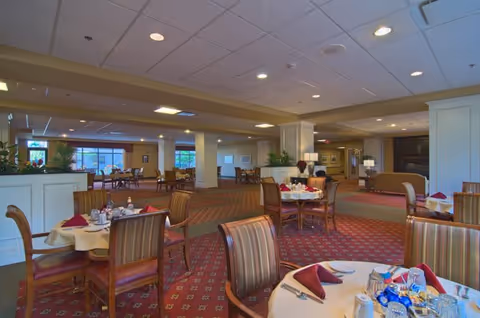 A spacious dining room with round tables covered in white tablecloths, set with plates, cups, and folded burgundy napkins. The room has patterned red carpet, wooden chairs with striped upholstery, and large windows letting in natural light. There are plants and decorative elements around the room, creating a warm and inviting atmosphere.