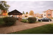 Outdoor courtyard at Elm Crest Senior Living Community with a wooden gazebo, walkway, shrubs, a parked car, and apartment buildings in the background.