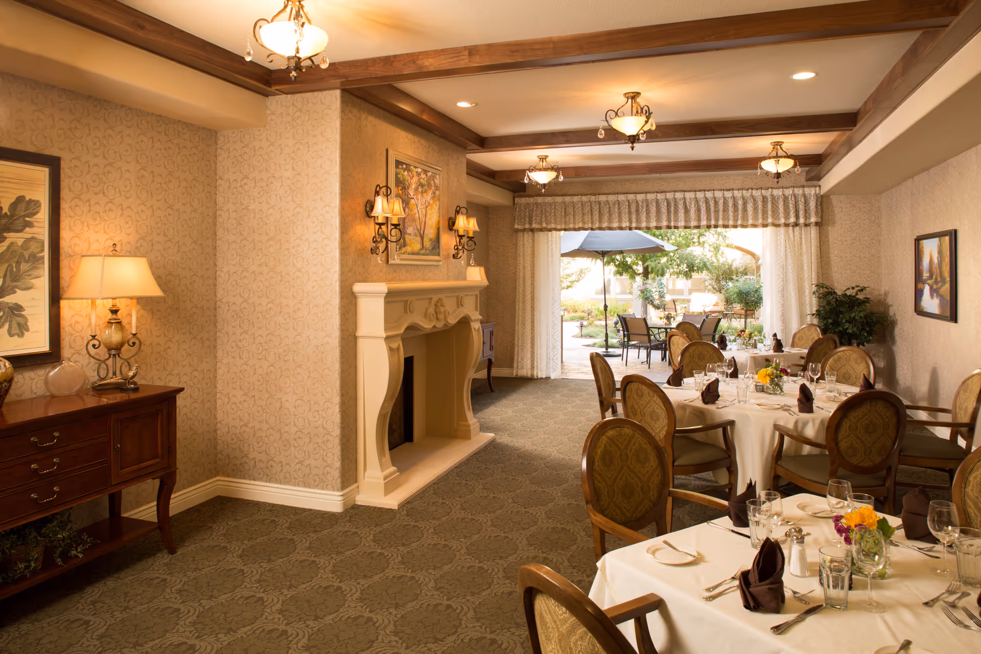Elegant dining room with round tables covered in white tablecloths, set with glassware, silverware, and folded napkins. The room features a decorative fireplace, wall sconces, framed artwork, and a side table with a lamp. Large windows with curtains open to an outdoor patio area with tables, chairs, and an umbrella surrounded by greenery.