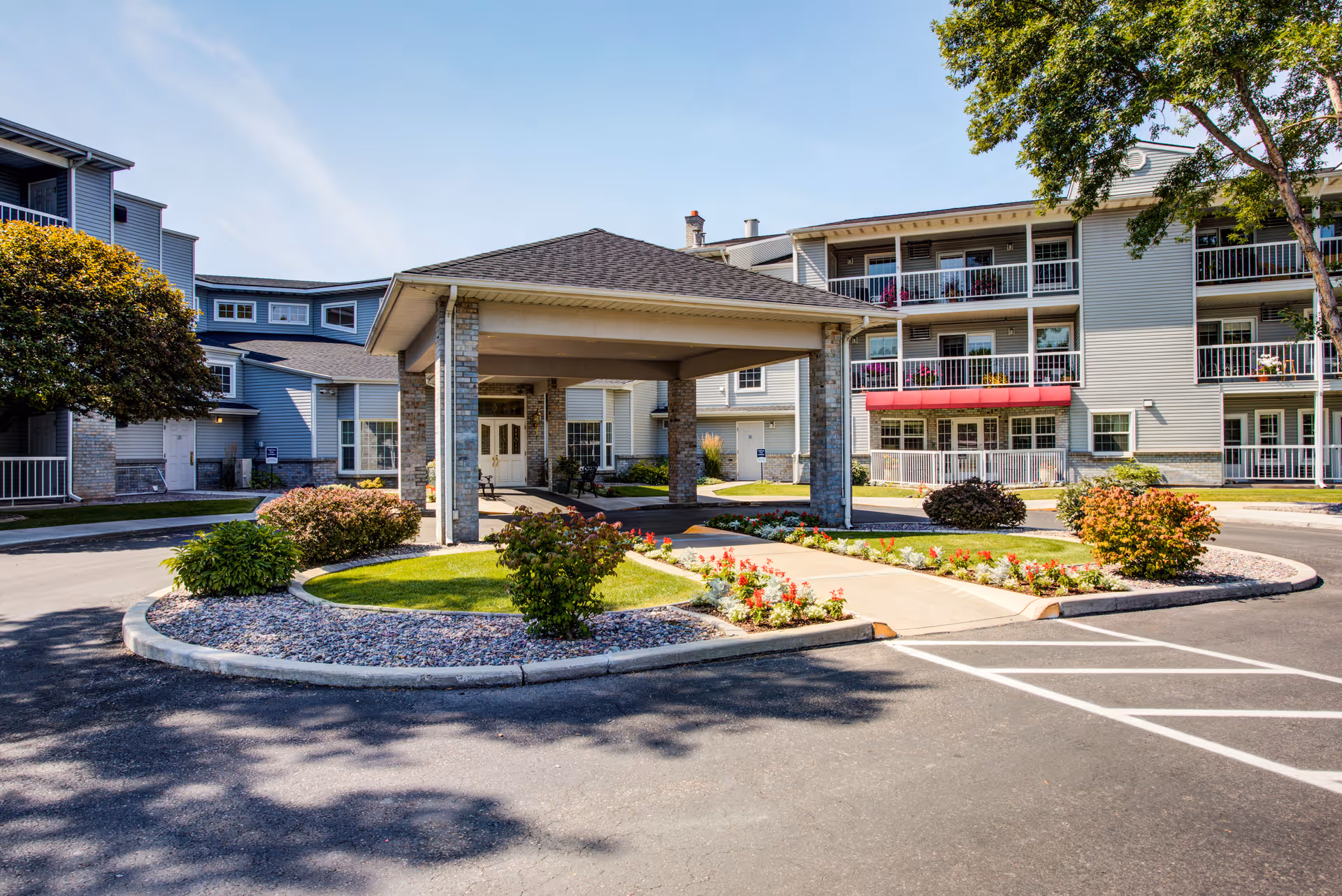 Entrance area of The Village Senior Residence featuring a covered drop-off zone with a circular driveway, landscaped flower beds, bushes, and a multi-story building with balconies in the background under a clear sky.