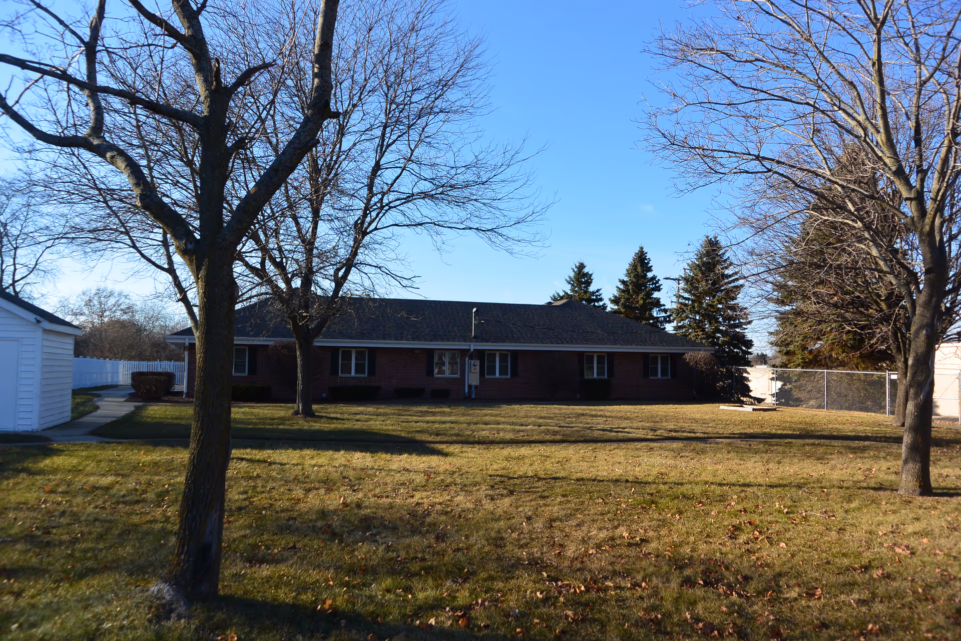 A single-story brick building with several windows, surrounded by a grassy yard with leafless trees under a clear blue sky.