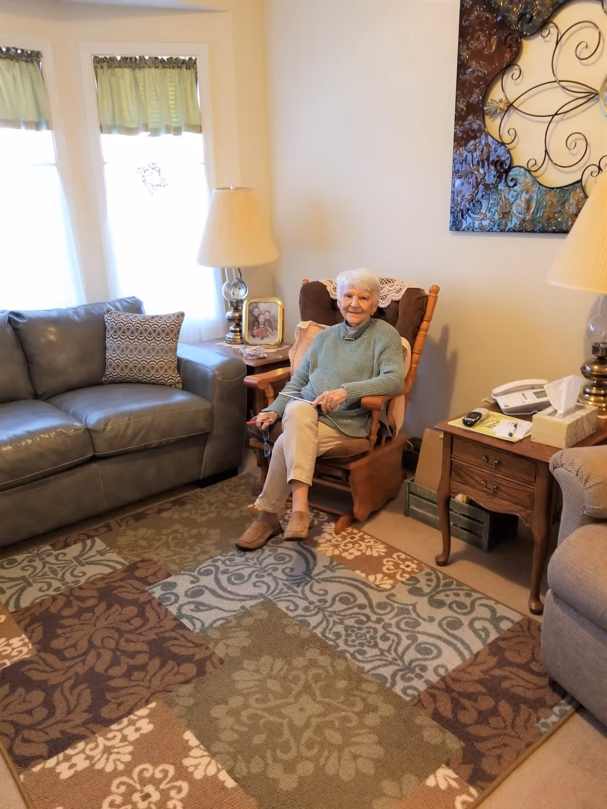An elderly woman with white hair wearing a green sweater and beige pants sits in a wooden rocking chair with cushions in a cozy living room. The room features a patterned area rug, a gray leather sofa with a decorative pillow, two table lamps on wooden side tables, a framed photo, a telephone, and a decorative wall hanging.