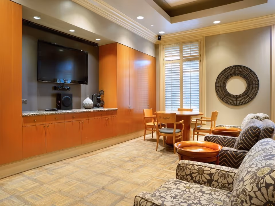 A cozy living room area with patterned armchairs and small wooden tables. There is a round wooden table with chairs near a window with white shutters. On the wall, there is a large flat-screen TV mounted above a granite countertop with wooden cabinets below. A decorative circular wall piece hangs on the adjacent wall.