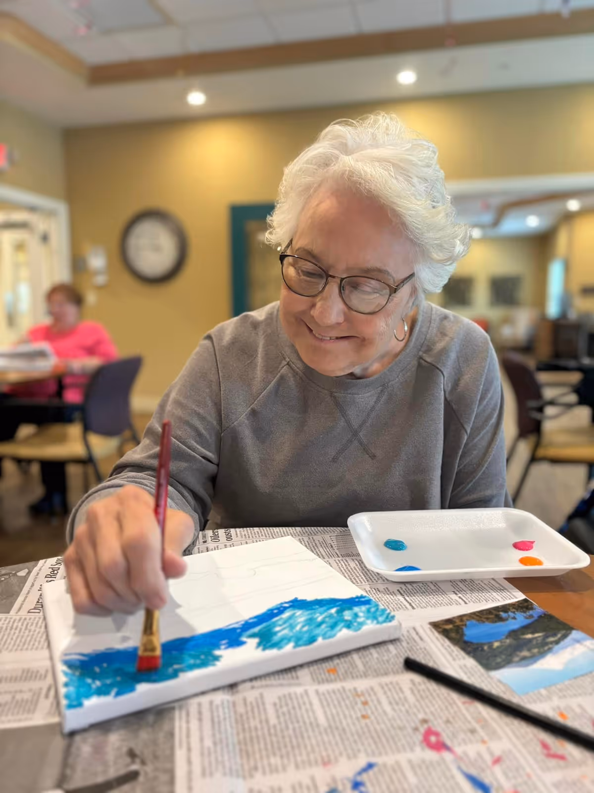 An elderly woman with white hair and glasses is smiling while painting on a canvas at a table covered with newspaper. She is using a paintbrush to apply blue paint. There are small blobs of paint on a white palette next to her. The background shows a room with other people and furniture, softly lit with warm tones.