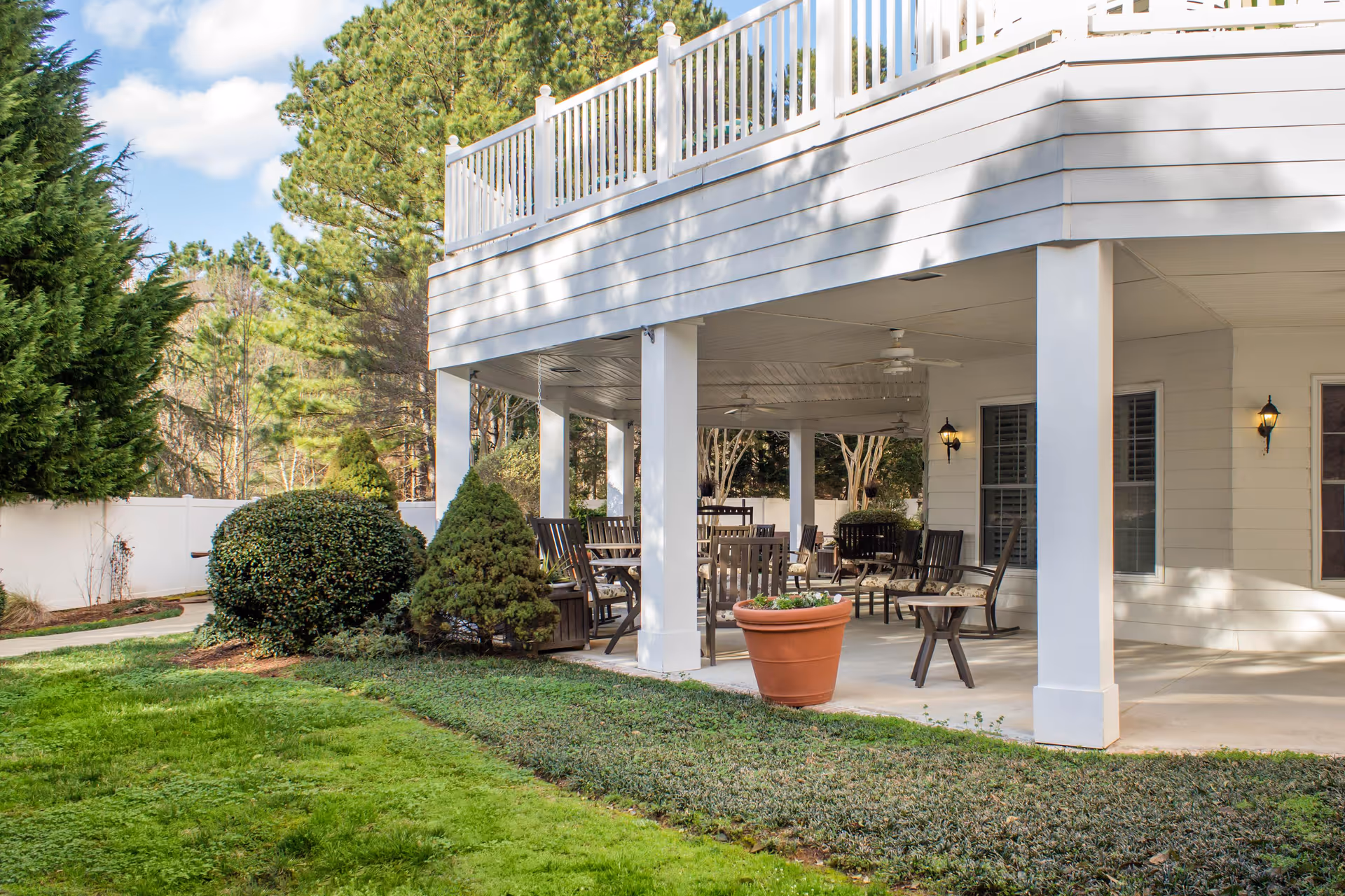Outdoor covered patio area with multiple wooden chairs and tables, ceiling fans, potted plants, and surrounding greenery including bushes and trees under a partly cloudy sky.