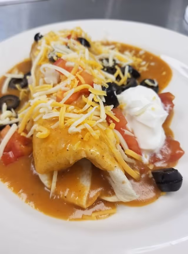 Close-up of a plate with saucy enchiladas topped with shredded cheese, diced tomatoes, sliced black olives and a dollop of sour cream.