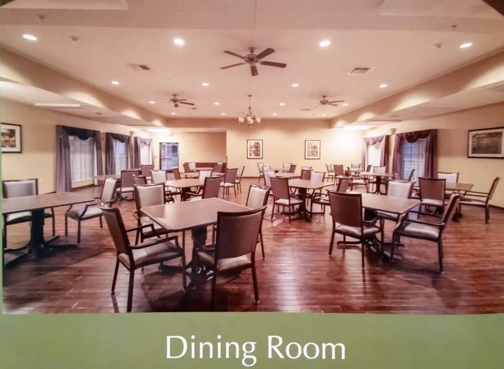A spacious dining room with multiple square tables and chairs arranged neatly on a wooden floor. The room has beige walls, several windows with curtains, ceiling fans, and framed pictures on the walls. The lighting is warm and recessed in the ceiling.