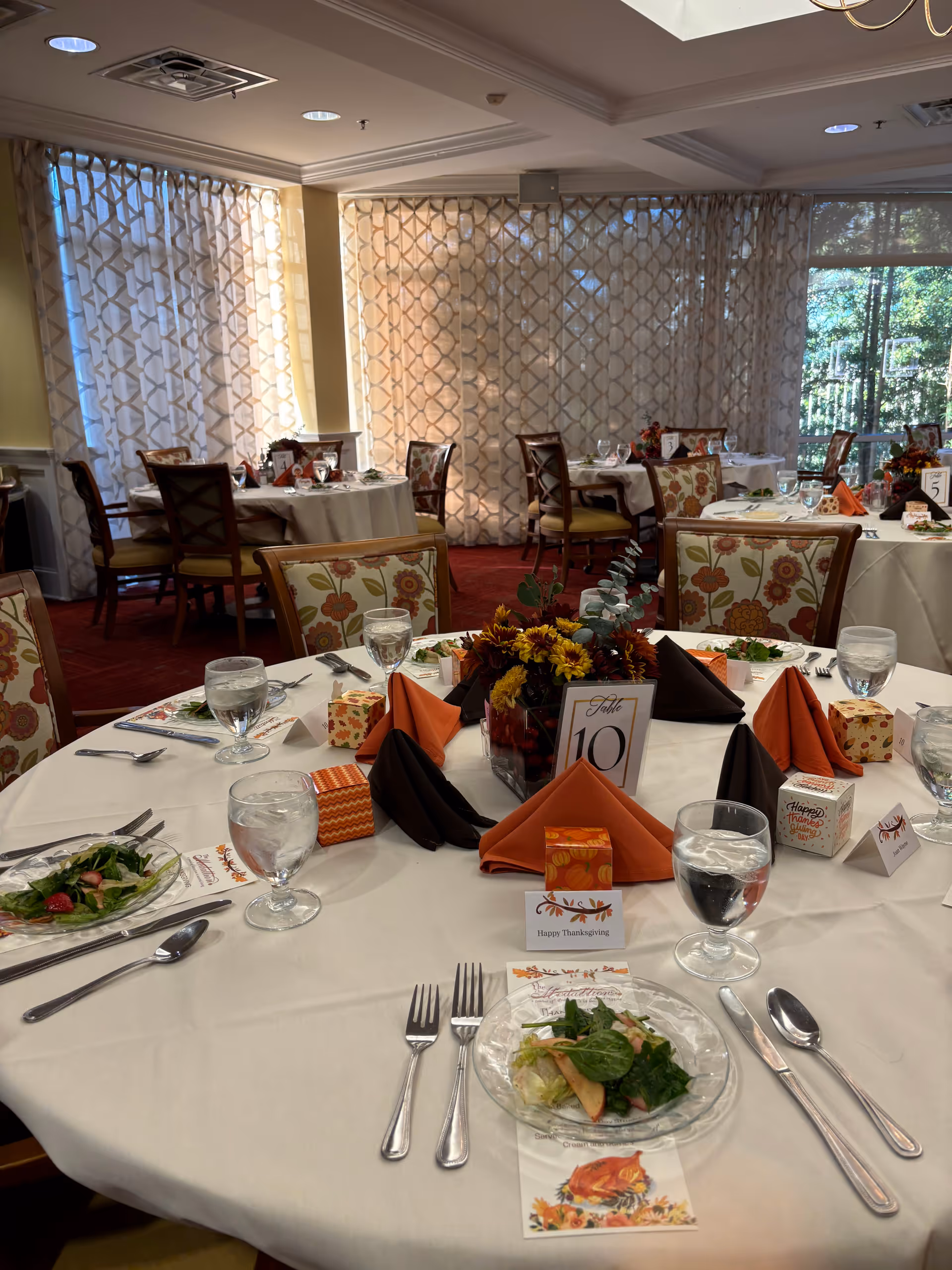 A dining room set up for a Thanksgiving meal with round tables covered in white tablecloths, each decorated with autumn-themed centerpieces, folded orange and brown napkins, place settings with plates of salad, glasses of water, and utensils. The room has patterned curtains and upholstered chairs with floral designs, and large windows letting in natural light.