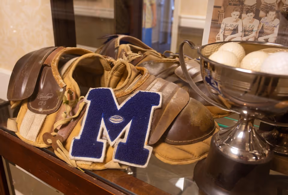 A display case containing vintage baseball equipment including a catcher's mitt, chest protector, and a blue letterman patch with the letter M. Next to the equipment is a silver trophy bowl filled with golf balls and a black and white photo of three young men in basketball uniforms.