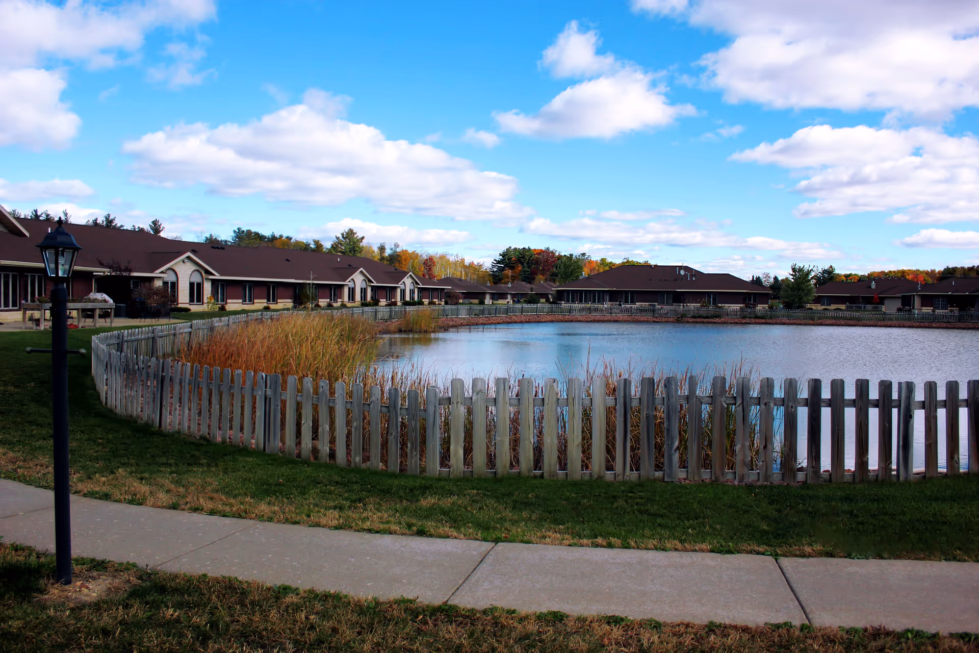 A scenic outdoor view of a senior living facility featuring a large pond surrounded by a wooden picket fence. The facility buildings with brown roofs and beige walls are visible in the background under a partly cloudy blue sky. There is a sidewalk and a black lamp post in the foreground.