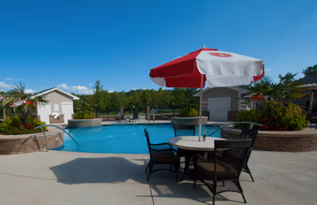 Outdoor swimming pool area with a round table and four chairs under a red and white umbrella. The pool is surrounded by small buildings, greenery, and a clear blue sky.