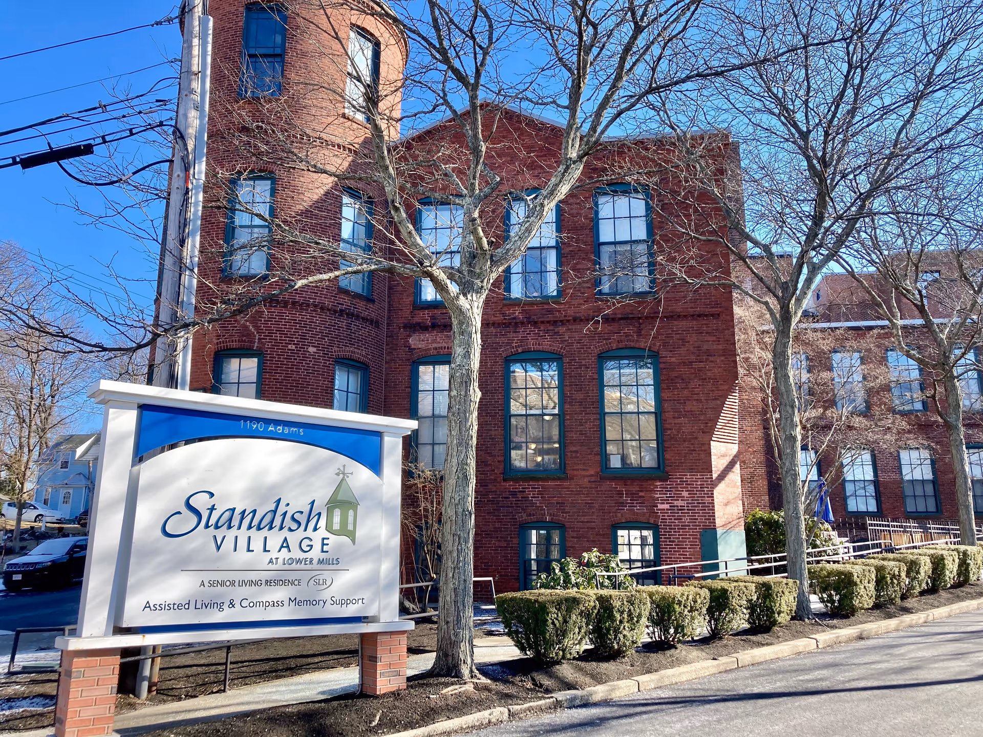 Exterior view of a red brick building with multiple windows and leafless trees in front. A large sign in the foreground reads 'Standish Village at Lower Mills, A Senior Living Residence, Assisted Living & Compass Memory Support'. The sky is clear and blue.