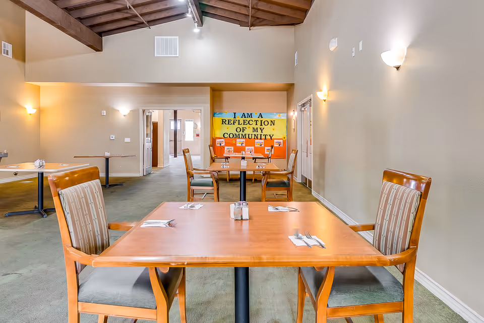 Dining room with wooden tables and chairs facing a colorful bulletin board that reads 'I AM A REFLECTION OF MY COMMUNITY'.