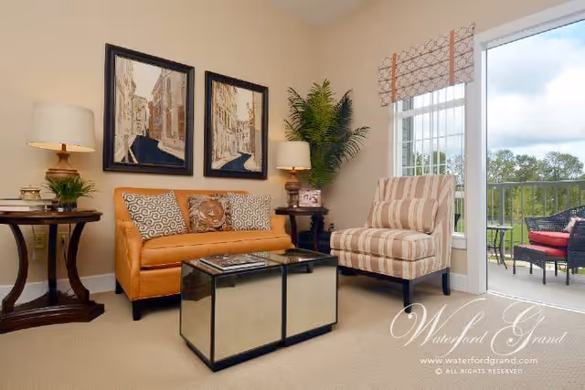 A cozy living room with a tan leather sofa adorned with patterned cushions, a striped armchair, two side tables each with a lamp and decorative items, a mirrored coffee table, two framed paintings on the wall, a large green plant, and a sliding glass door leading to an outdoor patio with wicker furniture and red cushions.