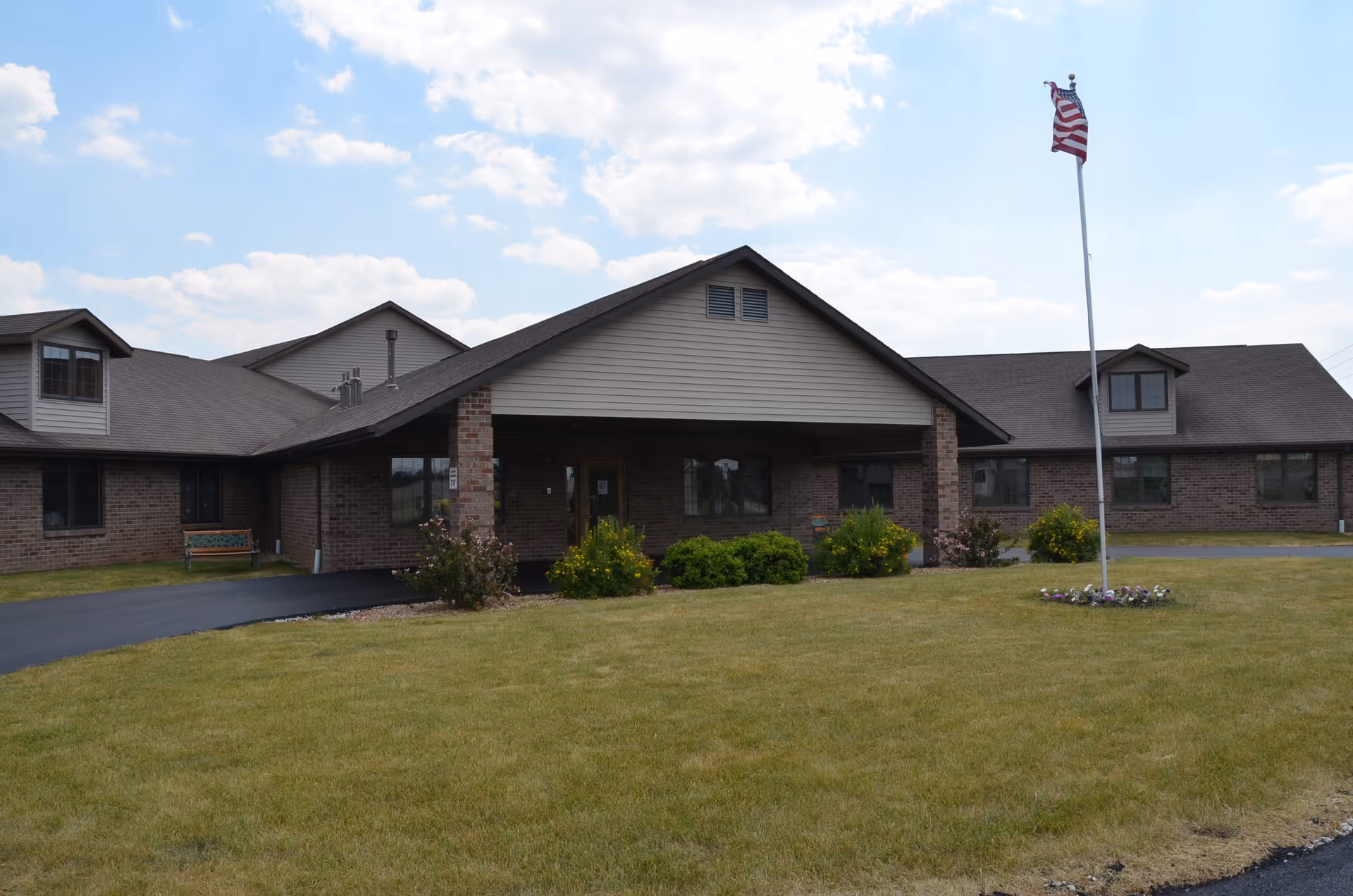 Exterior view of Century Ridge of Green Bay, a single-story brick building with a peaked roof and a covered entrance. There is a flagpole with an American flag in front of the building, surrounded by a small flower bed. The lawn is well-maintained with bushes and a bench near the entrance.