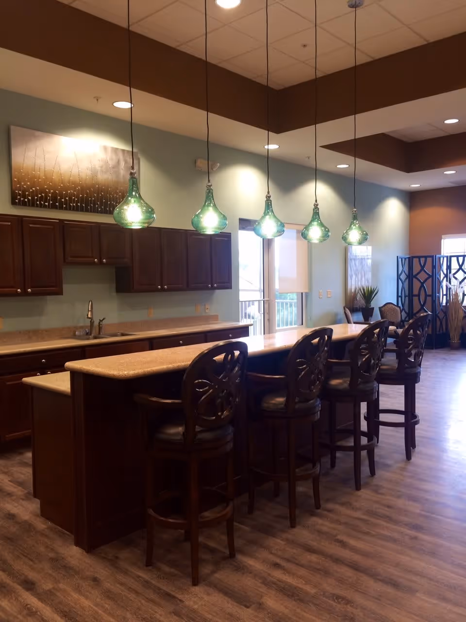 Interior view of a kitchen area with a long countertop island and five high-back bar stools. Above the island, five green pendant lights hang from the ceiling. The kitchen has wooden cabinets, a sink, and a light blue wall with a decorative painting. The floor is wood-patterned, and there is a window and some plants in the background.