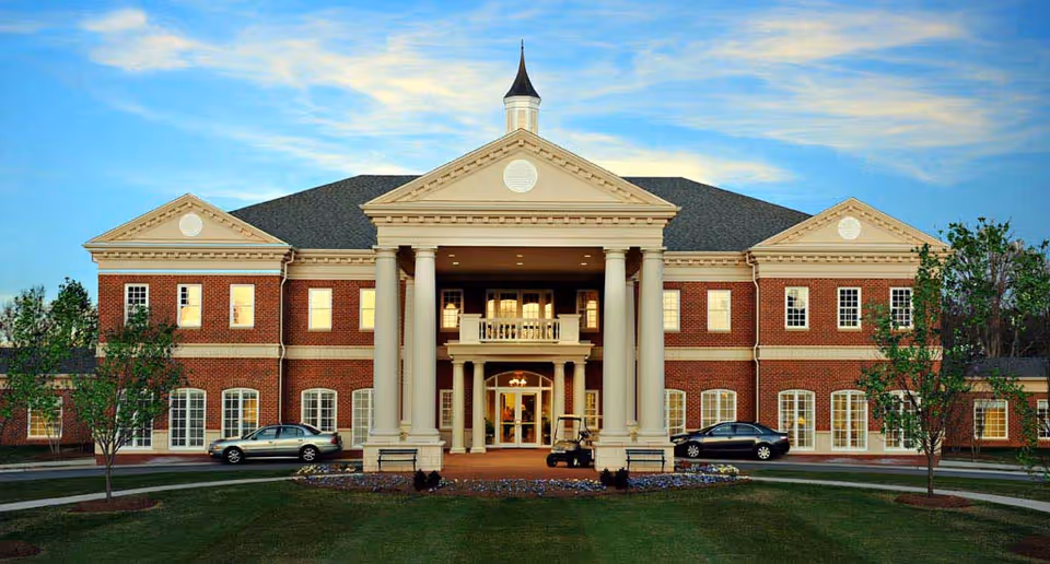 Front exterior view of a large, elegant brick building with white columns and a triangular pediment, surrounded by a well-maintained lawn and trees, under a partly cloudy sky at dusk.
