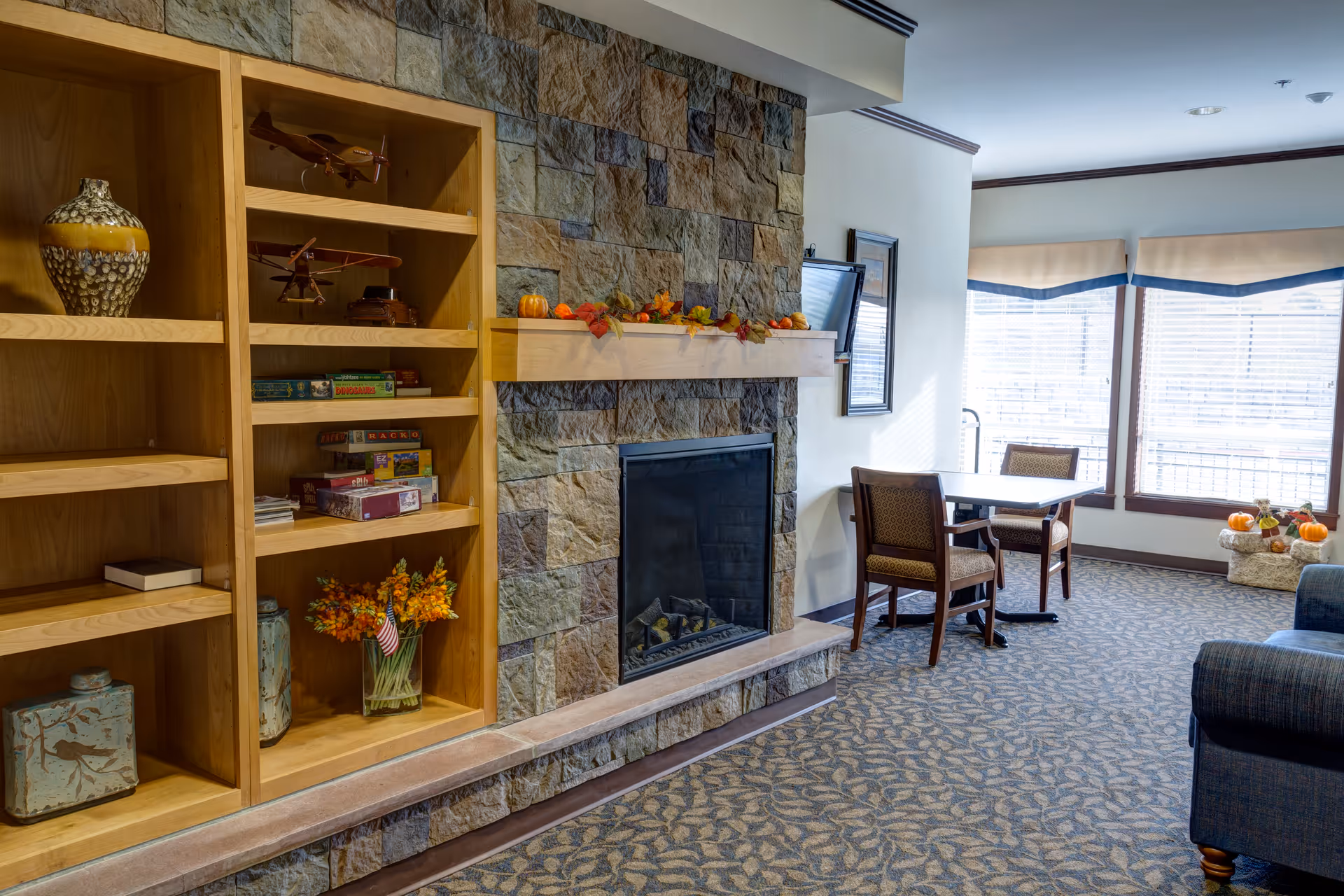 A cozy living room area featuring a stone fireplace with a wooden mantel decorated with autumn leaves and small pumpkins. To the left of the fireplace is a wooden built-in bookshelf with various decorative items, books, and board games. To the right, there is a small table with two chairs near large windows with blinds, allowing natural light to fill the room. The floor is carpeted with a patterned design, and part of a blue upholstered sofa is visible on the right edge of the image.