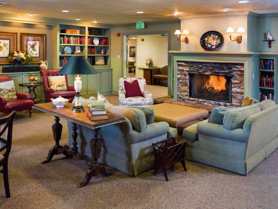 A cozy living room in a senior living facility featuring a stone fireplace with a fire burning, surrounded by green sofas and armchairs with floral and solid cushions. There is a wooden table with books and a lamp, built-in bookshelves filled with books and decorative plates, framed floral artwork on the walls, and soft lighting from wall sconces.